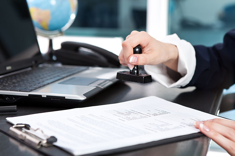 A person stamps a document on a clipboard at a desk with a laptop, phone, and globe in the background.