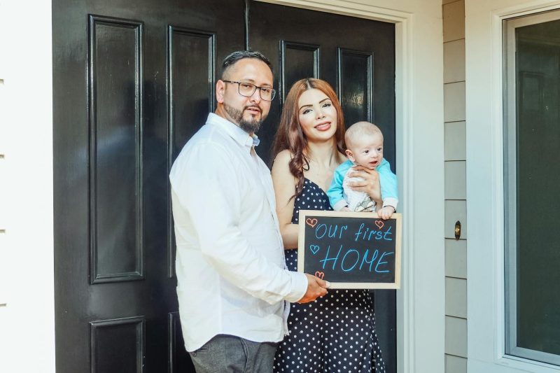 Young family with baby holding a sign Out First Home
