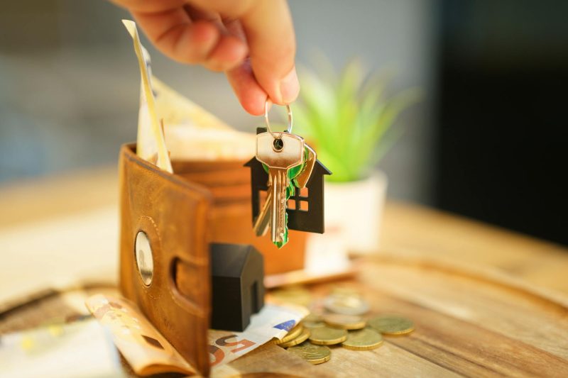 Hand holding a house key beside a wallet and coins on a surface.
