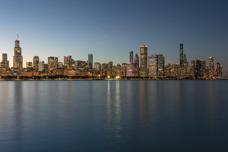 Chicago skyline at dusk with illuminated buildings reflected in a calm waterfront.