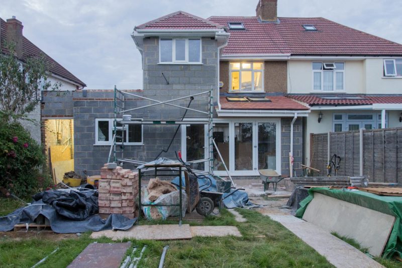 Suburban house under renovation with scaffolding, construction materials, and tarps in a residential neighborhood.