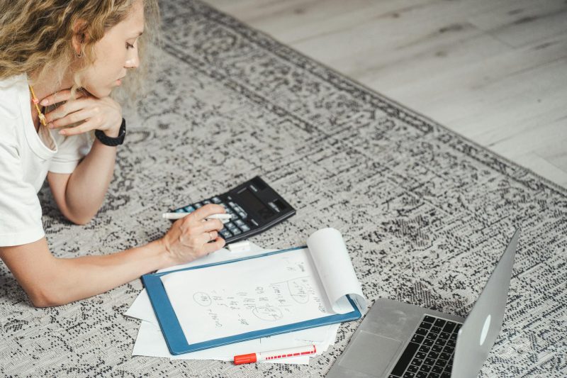Woman sitting on a rug, using a laptop and calculator while reviewing financial documents.